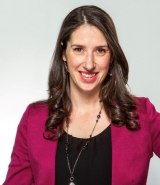 A woman with long, dark hair is smiling at the camera. She wears a magenta blazer over a black top, accessorized with a long necklace. The background is a light neutral color, making her stand out prominently. She exudes a friendly and professional demeanor.