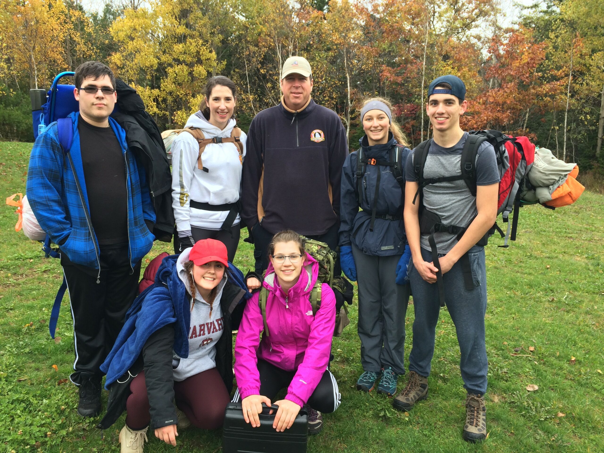 A group of seven people are posed together, looking at the camera. They're all dressed in warm, casual clothing with backpacks and gear. The background features vibrant autumn trees with yellow, orange, and red leaves. Five people are standing in the back wearing hoodies, jackets, and hiking attire. Two women kneeling in front, one in a red cap and the other in a bright pink jacket.