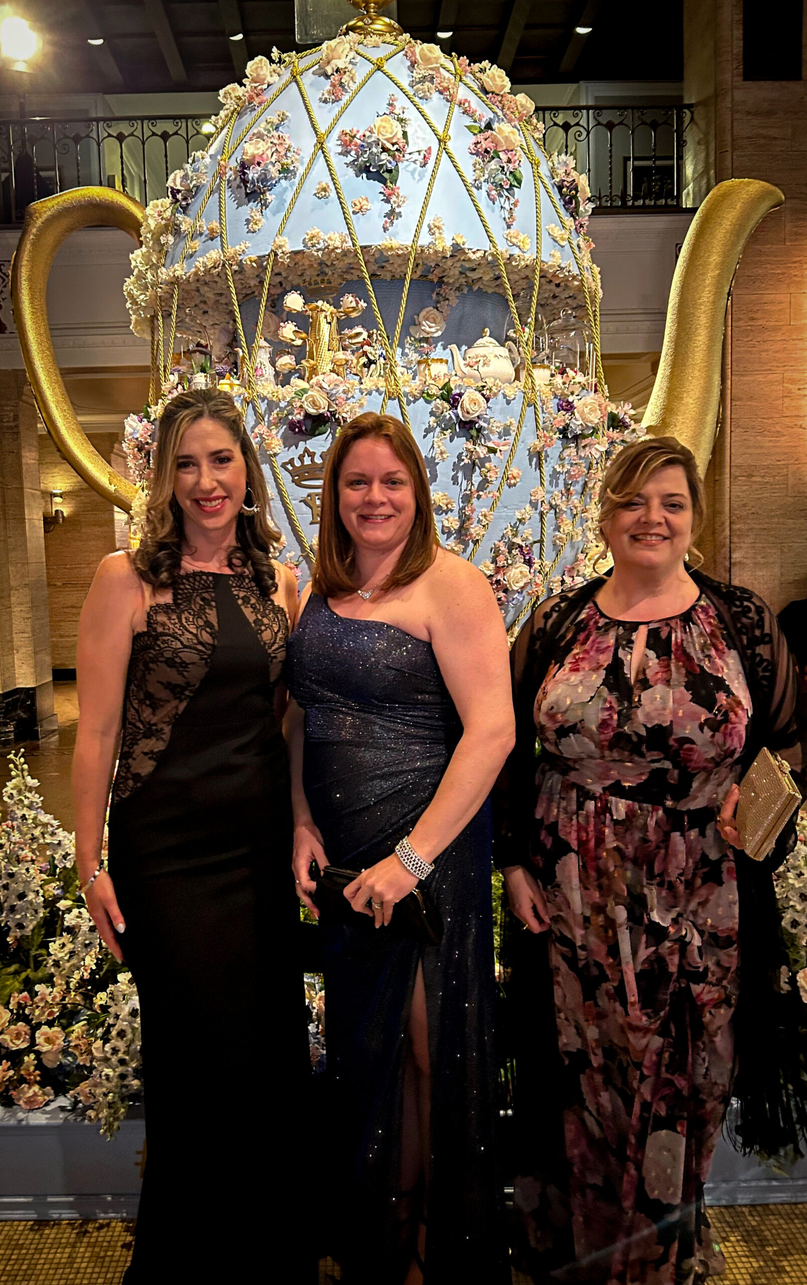 Three women dressed in elegant evening gowns pose in front of a large, decorative teapot adorned with intricate floral designs and gold accents. The woman on the left wears a black gown with lace details, the woman in the middle is in a sparkling navy blue one-shoulder dress, and the woman on the right is wearing a floral-patterned gown with black lace. They all smile warmly at the camera while holding small clutches. The background features soft lighting, enhancing the grand and luxurious atmosphere.