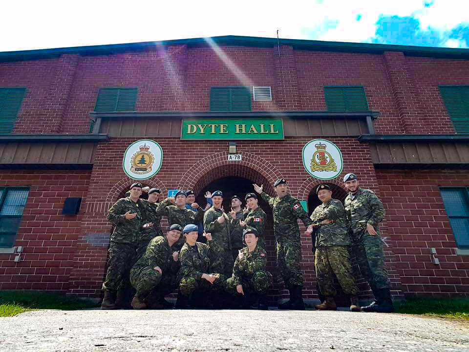 A group of cadets wearing army uniforms cheerfully posing in front of a red brick building. Most of them are standing with three squatting low in the front row. A sign above the archway behind them reads Dyte Hall.