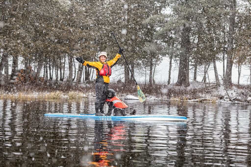 A person dressed in winter gear, including a yellow jacket, red life vest, and knit hat, stands on a paddleboard in the middle of a calm lake while it is snowing. They are holding a paddle with one hand and have their other arm raised. A black dog wearing an orange life vest sits on the paddleboard in front of them. Snow-covered trees line the background, and large snowflakes are visible in the air, creating a serene winter scene.