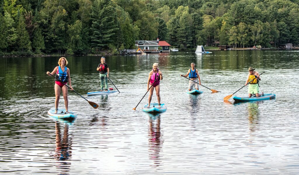 Five people paddleboarding on a calm lake surrounded by dense green trees. They are evenly spaced, each standing on their own paddleboard and holding a paddle. Two women in bright swimsuits and life vests are in the front and two men in casual outdoor clothing and life vests are on the sides, with the teacher in the back. The reflections of the paddleboarders are visible on the still water. In the background, there is a lakeside cabin with a dock, along with some floating water inflatables. The scene is serene and captures the relaxed atmosphere of a summer day on the water.