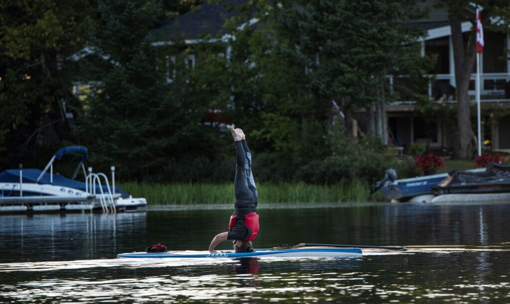 A woman wearing a red life vest, performs a headstand on a paddleboard, showcasing balance and skill. The water is calm and flat.