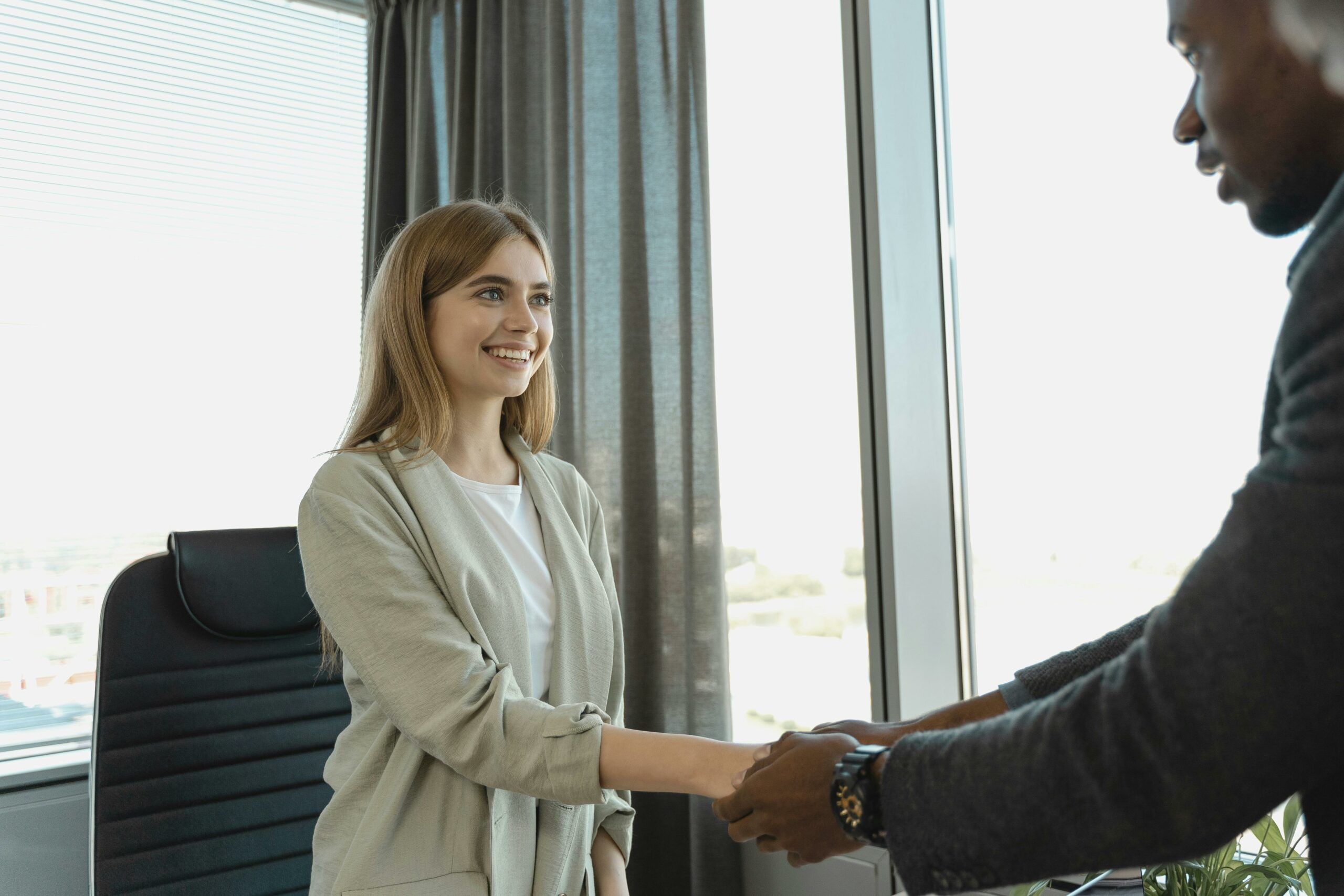 A female job candidate with blonde hair shakes hands with the interviewer. She is smiling brightly.