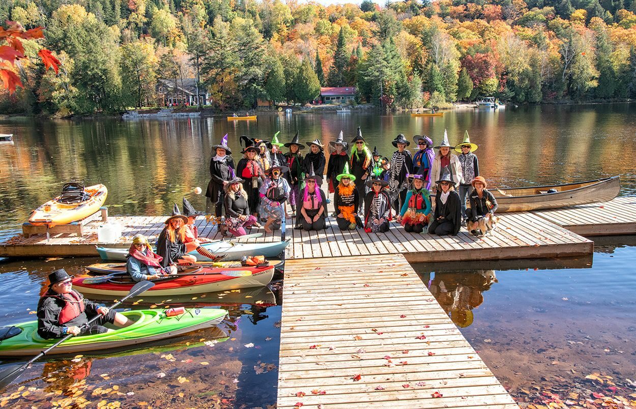 A group of people, many dressed in colorful Halloween costumes, stand on a wooden dock by a scenic lake. Some are wearing witch hats, capes, and other festive attire. Four kayakers, also in costume, sit in their kayaks on the water near the dock. In the background, there are vibrant autumn trees, with leaves in shades of orange, yellow, and red. The calm lake reflects the trees and costumes, creating a picturesque, fun, and seasonal atmosphere.